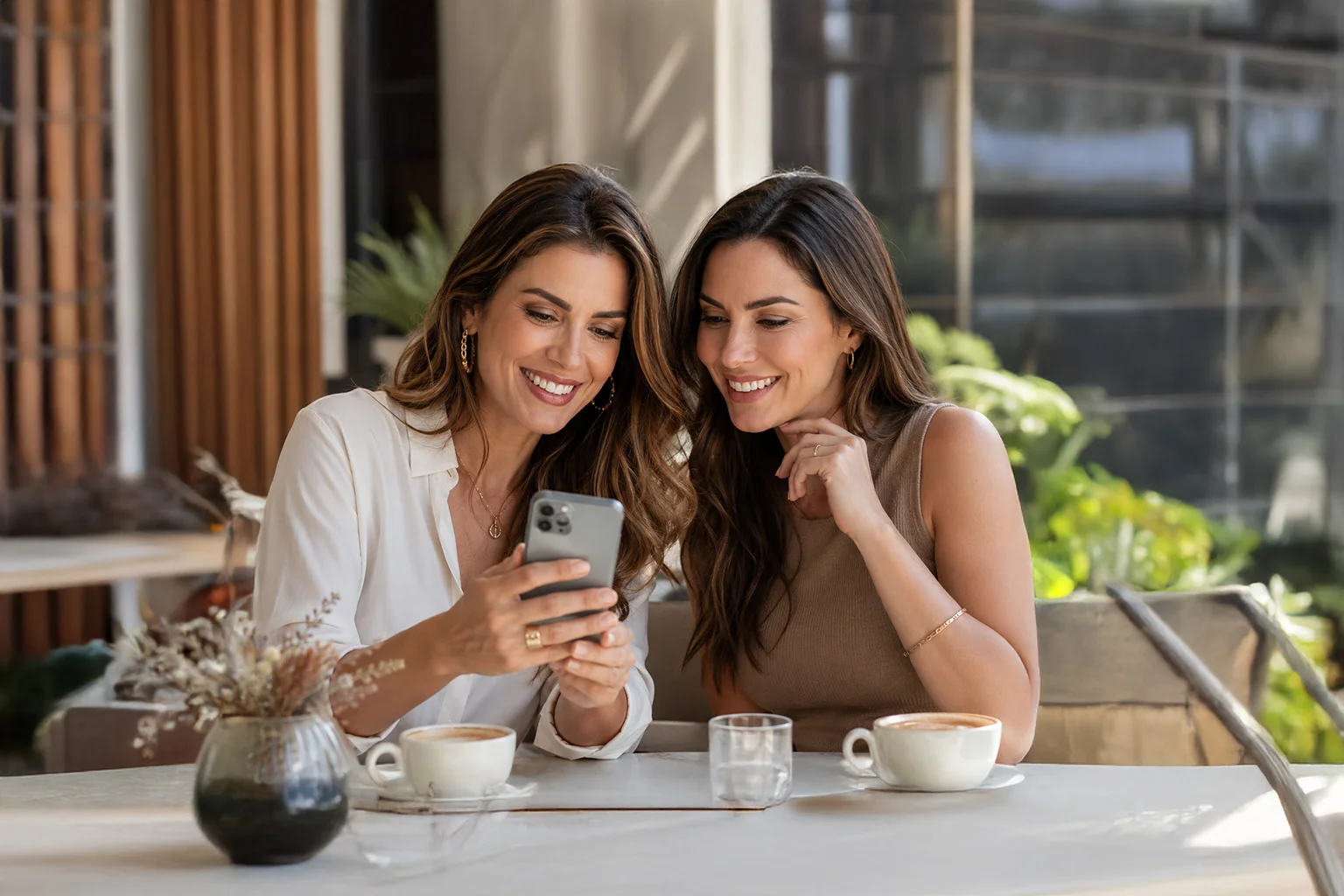 Duas amigas sorrindo enquanto olham o celular juntas em um café