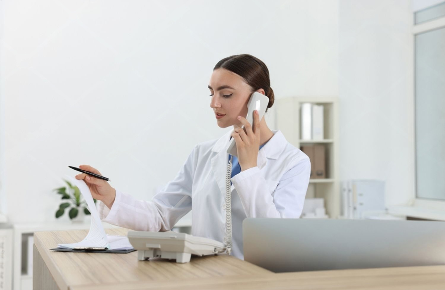 Profissional de jaleco ao telefone, com papéis e computador sobre a mesa